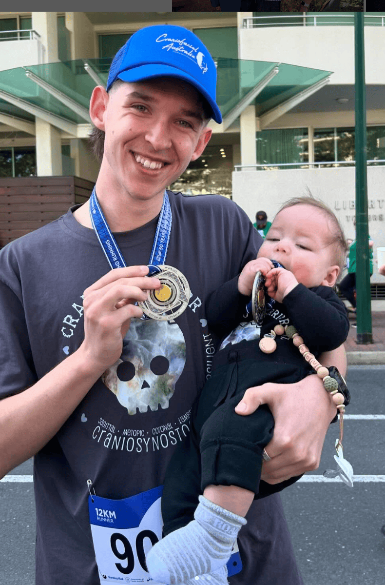 Baby with sagittal synostosis held by his uncle, who is wearing a Cranio Warrior t-shirt and holding a medal