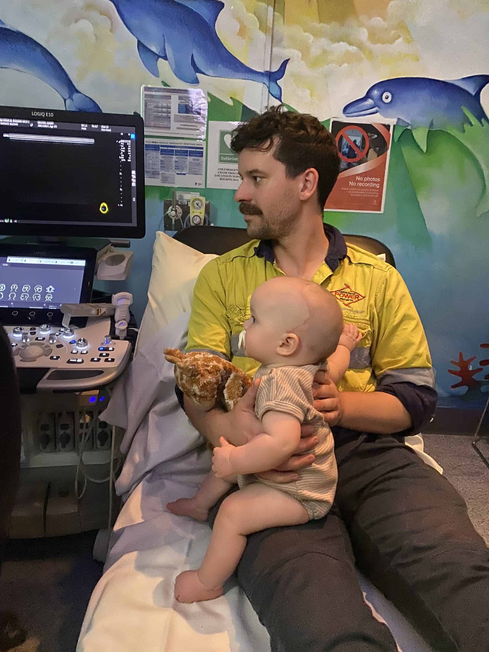 Baby with sagittal synostosis held by his father following craniofacial surgery, sitting on a hospital bed