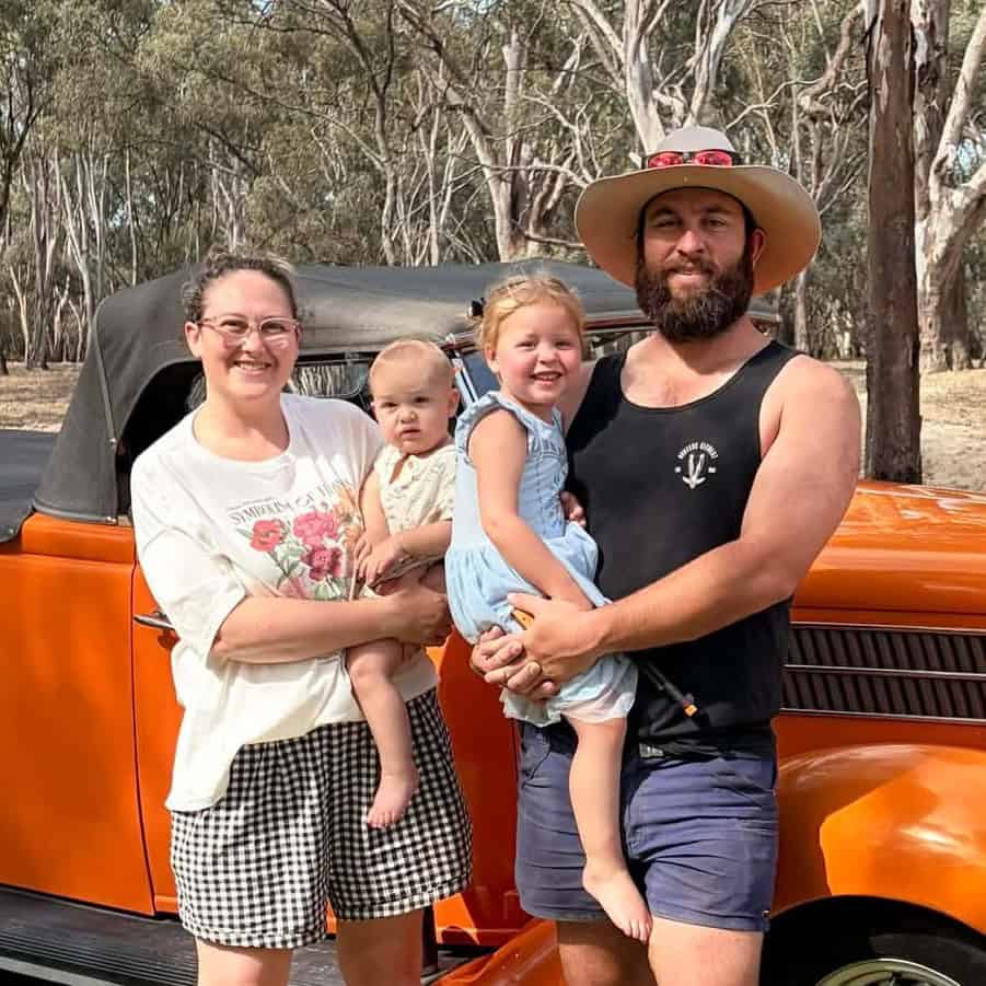 A family with two children standing outdoors next to an orange vintage vehicle, smiling.