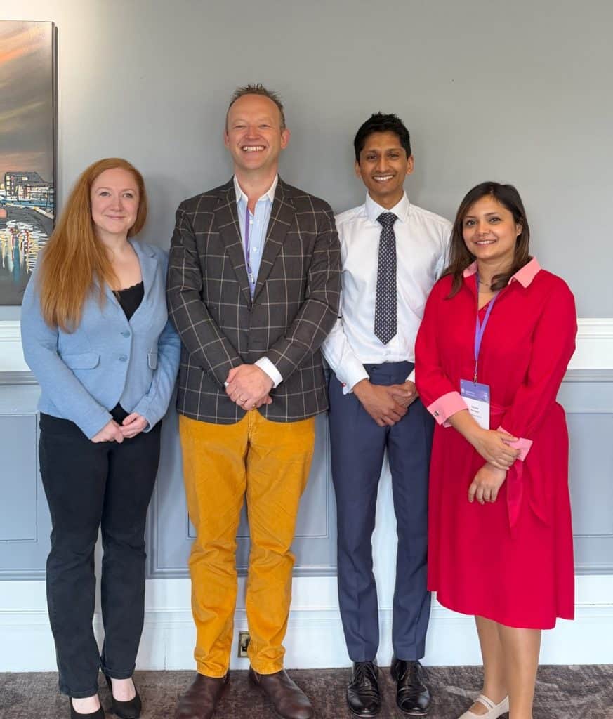 A group of four diverse professionals smiling at a conference or seminar, standing in front of a gre.