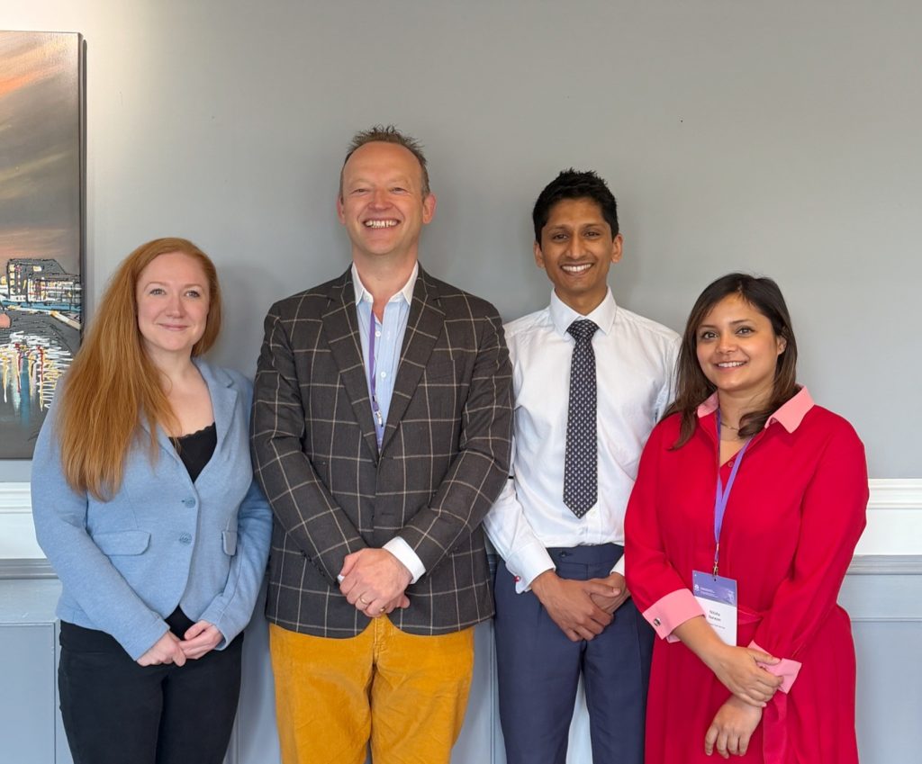A group of four diverse professionals smiling at a conference or seminar, standing in front of a gre.