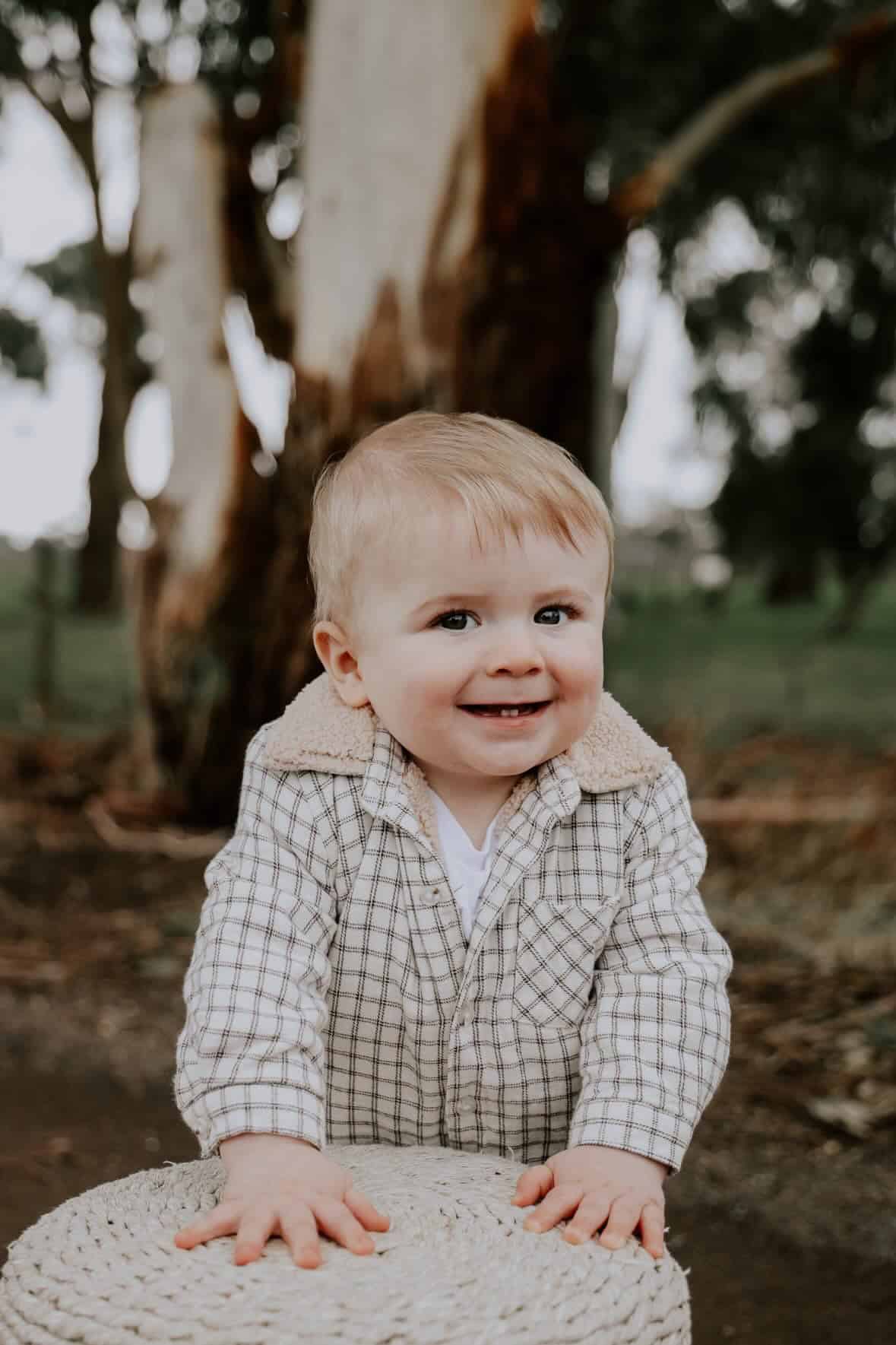 Smiling toddler outdoors near a tree in a checkered shirt.