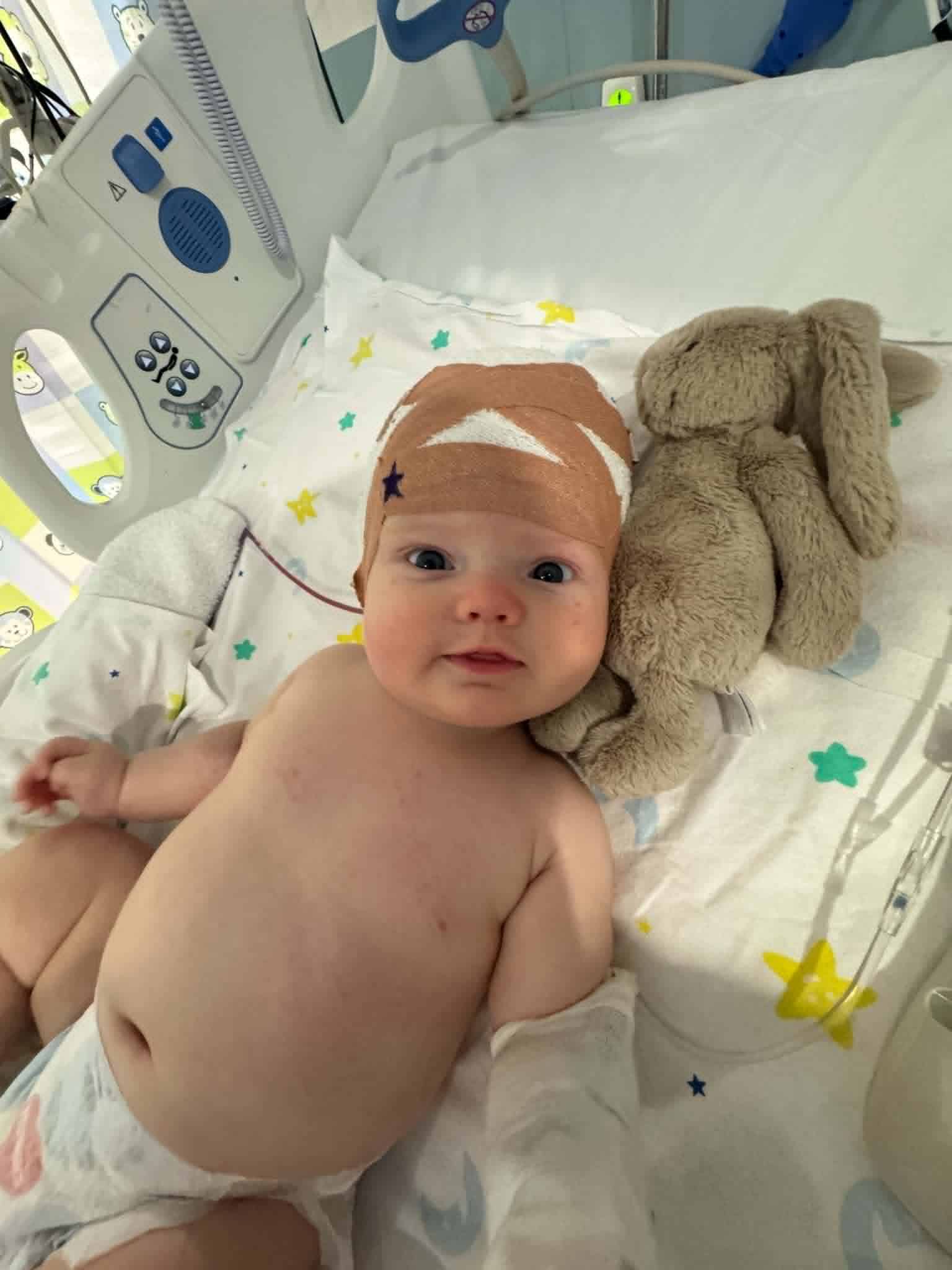 Baby in hospital bed with bandage and teddy bear, medical equipment nearby.