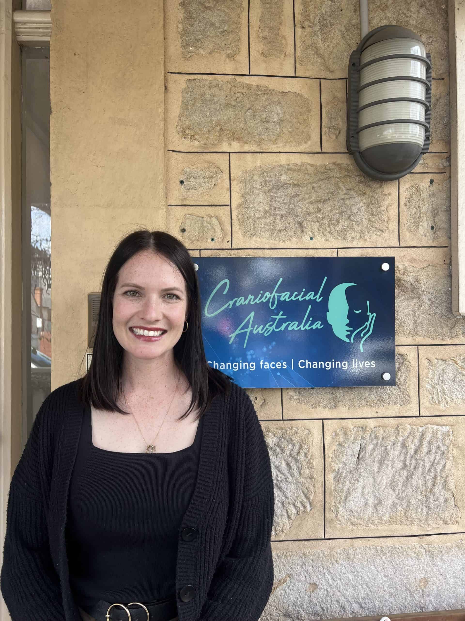 Smiling woman standing outside Craniofacial Australia clinic with sign and stone wall.