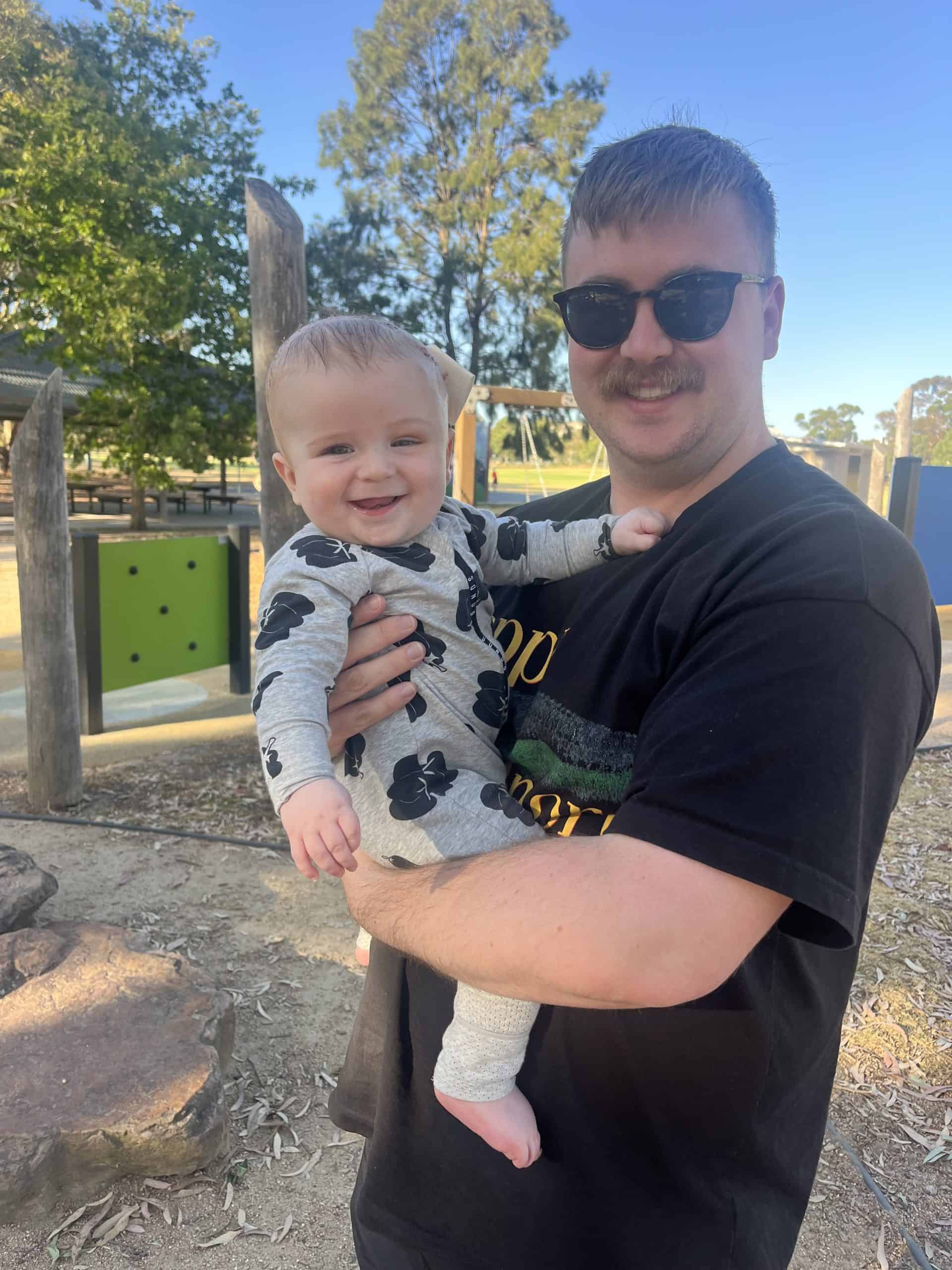 Man holding smiling young girl outdoors in park with trees and playground equipment.