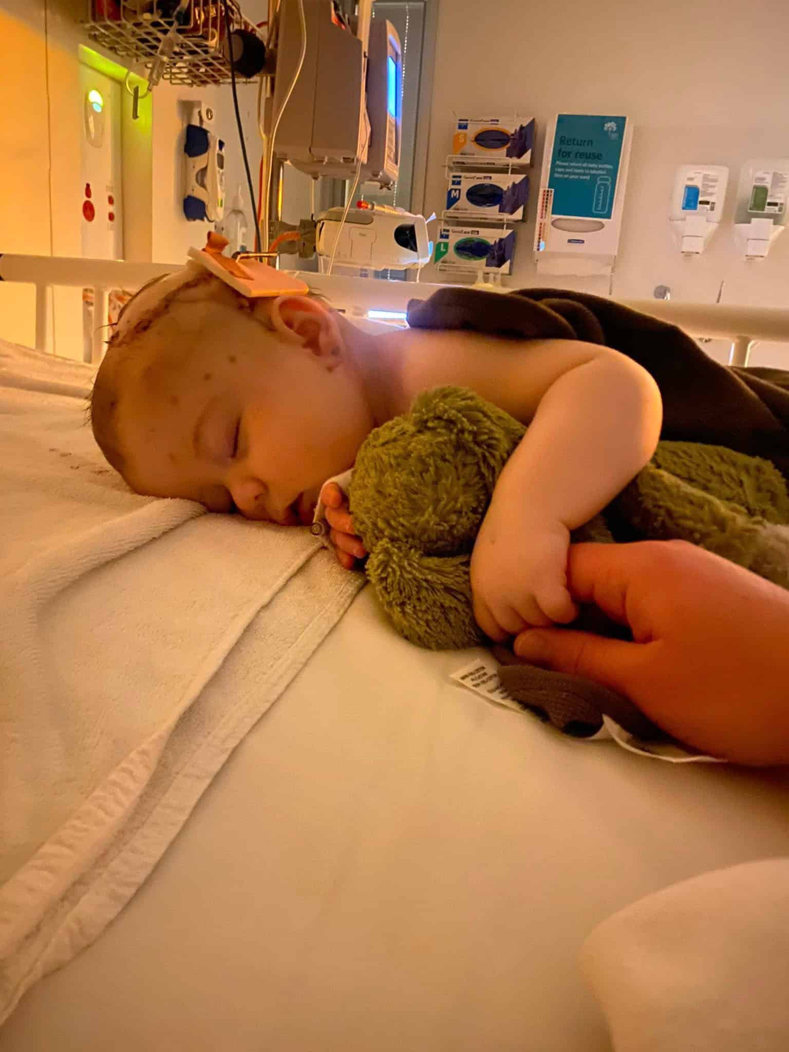 Young child sleeping in hospital bed holding a green teddy bear.