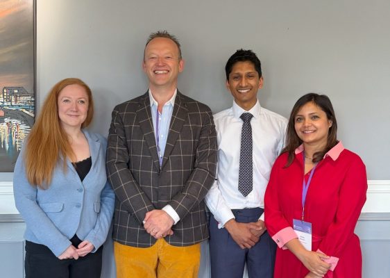 A group of four diverse professionals smiling at a conference or seminar, standing in front of a gre.